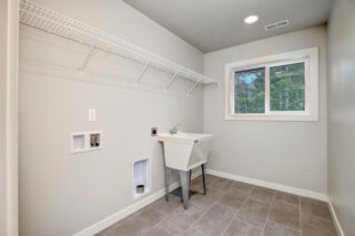 A tidy laundry room with a washing basin, a metal shelf, a window with a forest view, tiled flooring, and a pet door built into the wall.