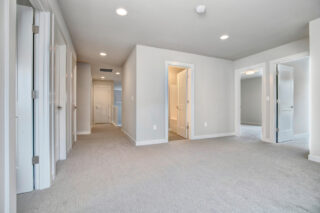 The image shows a bright, empty room with white walls and carpet flooring. Multiple doors suggest a layout with several rooms or closets.