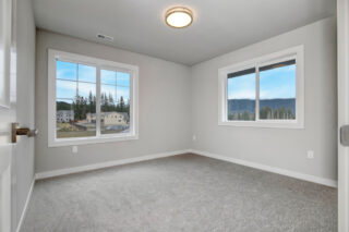 An empty room with gray walls and carpet, two windows offering views of a residential area and mountains, and a ceiling light fixture.