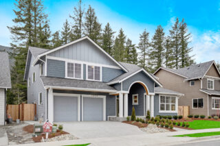 This is a suburban neighborhood featuring new two-story houses with an arched entryway, a double garage door, and a for-sale sign. Tall trees and cloudy sky background.