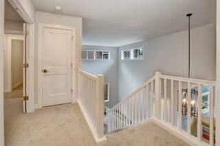 A well-lit, neutral-toned hallway with carpeted floors, white railings, a closed door, an open door, and a modern hanging light fixture.