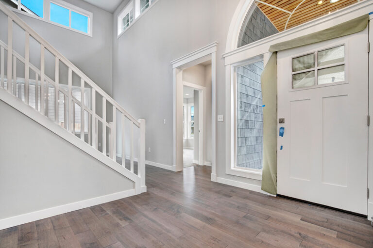 An empty foyer with high ceilings, a staircase, wood floors, and a white front door. The room features modern architecture and ample natural light.