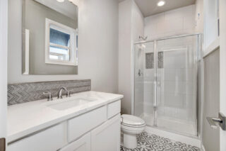 A modern bathroom with a white vanity, a large mirror, a glass-walled shower, patterned floor tiles, and a small window allowing natural light.