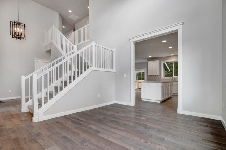 The image shows a modern interior with a wooden floor, white staircase, and a glimpse into a kitchen with cabinets and tiled backsplash.