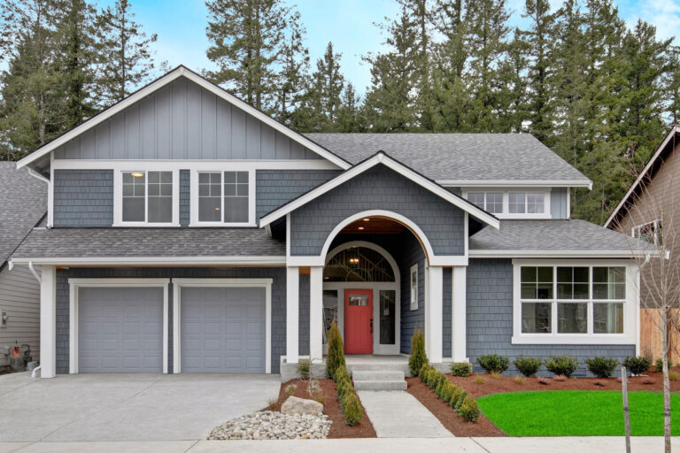 A two-story suburban house with gray siding, a red door, dual garage doors, lush green lawn, and trees in the background on an overcast day.