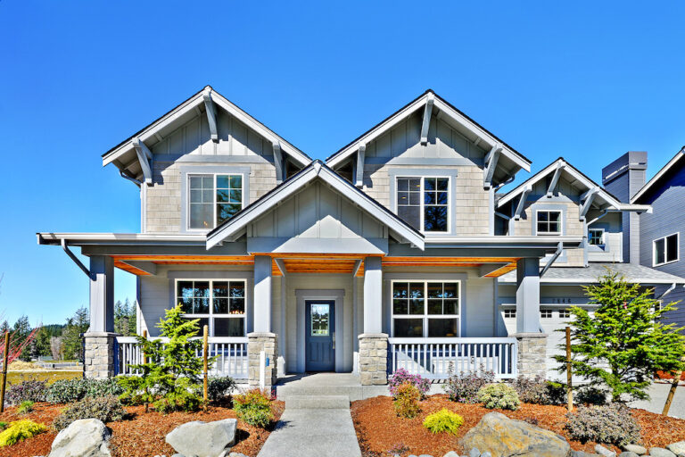  A two-story house with a gable roof, stone columned porch, large windows, landscaped front yard, and a clear blue sky background. 