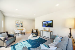 This image displays a bright, modern living room with a gray sectional sofa, two leather chairs, a round dining set, wall art, and a television.