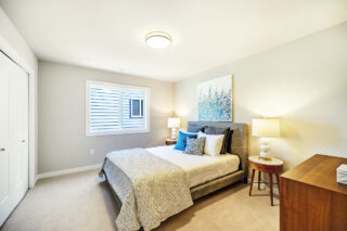 A bright, modern bedroom with a large bed, white and blue bedding, two nightstands with lamps, artwork above the bed, and neutral walls.