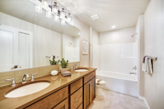 Modern bathroom with double vanity, large mirror, wooden cabinets, white walls, tiled floor, bathtub, toilet, and hanging lights. Clean and well-lit.