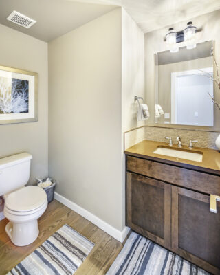 The image shows a modern bathroom interior with a toilet, a wooden vanity with sink, a mirror with lights, and a decorative rug on the floor.