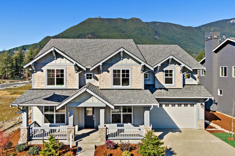 A two-story suburban house with a gray shingle roof, beige siding, a two-car garage, and a landscaped yard, set against a mountainous backdrop.