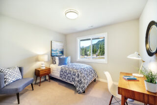 This is a bright, modern bedroom with a patterned bedspread, a desk, and a window showing a mountainous landscape. The room has contemporary furnishings and artwork.