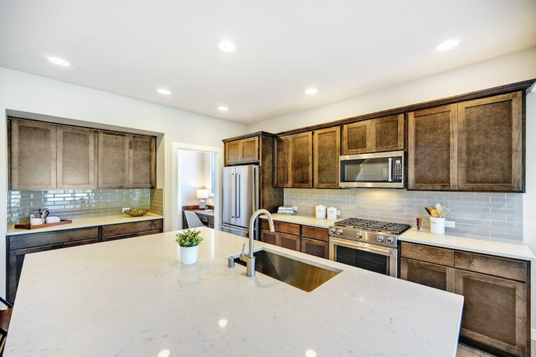 A modern kitchen with dark brown cabinets, stainless steel appliances, a white marble countertop island, and a subway tile backsplash.