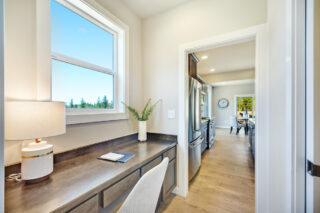 A bright, modern home interior with a desk, lamp, and plant by a window overlooking trees. An open doorway leads to a kitchen and dining area.