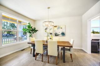 A bright, modern dining room featuring a wooden table with cream chairs, contemporary art, a large window with a forest view, and a pendant light.