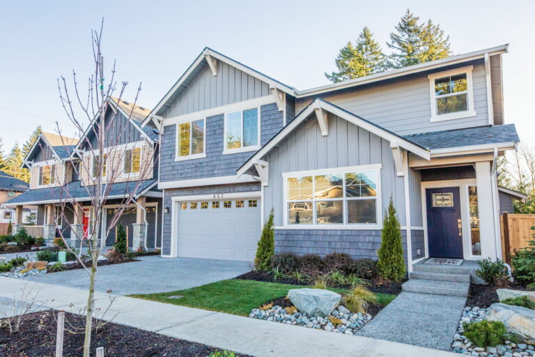  This is a two-story suburban house with gray siding, white trim, a garage, and a landscaped yard with a young leafless tree. 