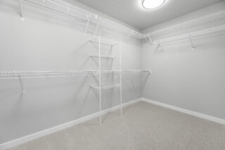 An empty walk-in closet with white wire shelving on light gray walls, featuring a carpeted floor and a central ceiling light.