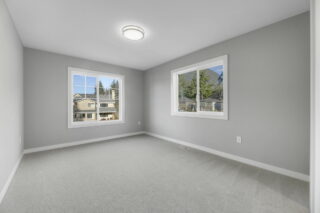 Empty room with light gray walls, carpeted floor, two windows with a view of neighboring houses, and a central ceiling light fixture.