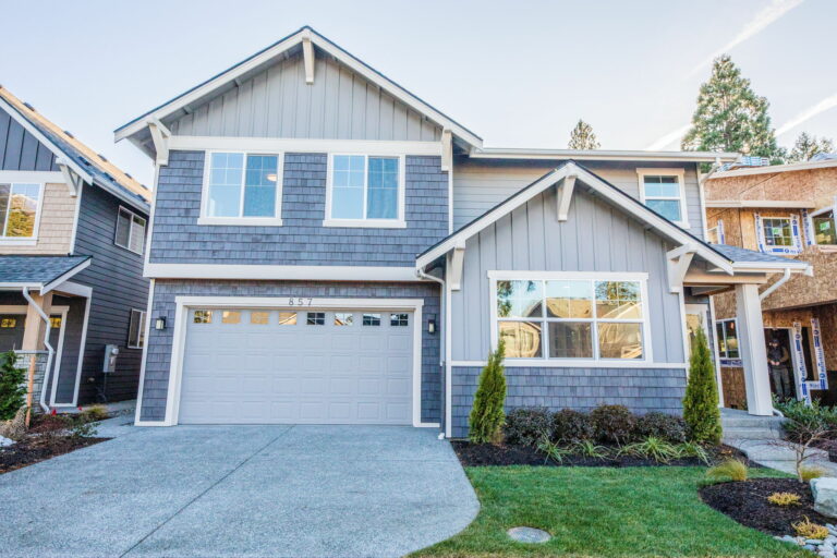 A modern two-story house with gray shingles, white trim, a double garage door, a well-manicured lawn, and green shrubbery under a clear sky.