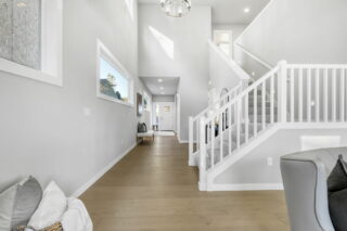 A bright, spacious hallway with hardwood floors, a white staircase, framed art on the walls, and modern lighting fixtures. There's minimalist furniture present.