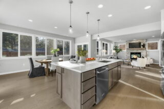 A modern open-plan kitchen and living area with light wood floors, white countertops, stainless steel appliances, and a stone fireplace. Large windows provide natural light.