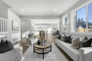 Modern open-plan living room with neutral tones, large windows letting in natural light, contemporary furniture, leading to a dining area and kitchen.