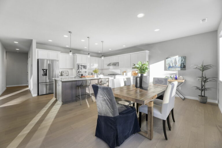 Modern kitchen with white cabinets, stainless steel appliances, and an island. Open-plan to a dining area with a wooden table and white chairs. Bright, minimalist design.