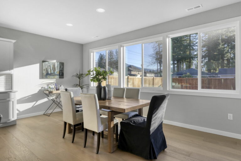 A modern dining room with a wooden table, white chairs, large windows showing trees outside, hardwood floors, and muted decor.