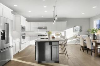 A modern kitchen with white cabinetry, a central island, stainless steel appliances, and a dining area with a wooden table and chairs bathed in sunlight.