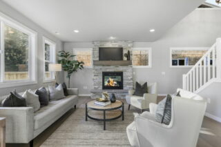 A modern living room with a stone fireplace, mounted TV, neutral tone sofas, decorative pillows, round coffee table, windows, and a staircase.