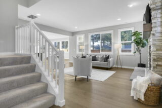 An elegant living room with natural light, modern furniture, neutral colors, a stone fireplace, and wooden flooring. Stairs lead up to another level.