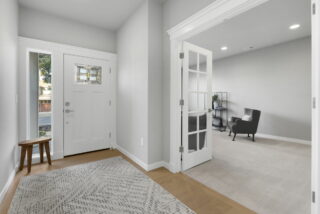 Modern entryway with white door, stained glass, a wooden bench, patterned rug, and adjacent room with armchair, shelving unit, and French doors.