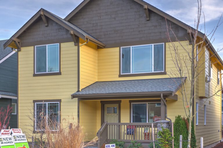 A two-story yellow and gray house with a "New Homes" sign, landscaped front yard, porch, and clear skies in the background.