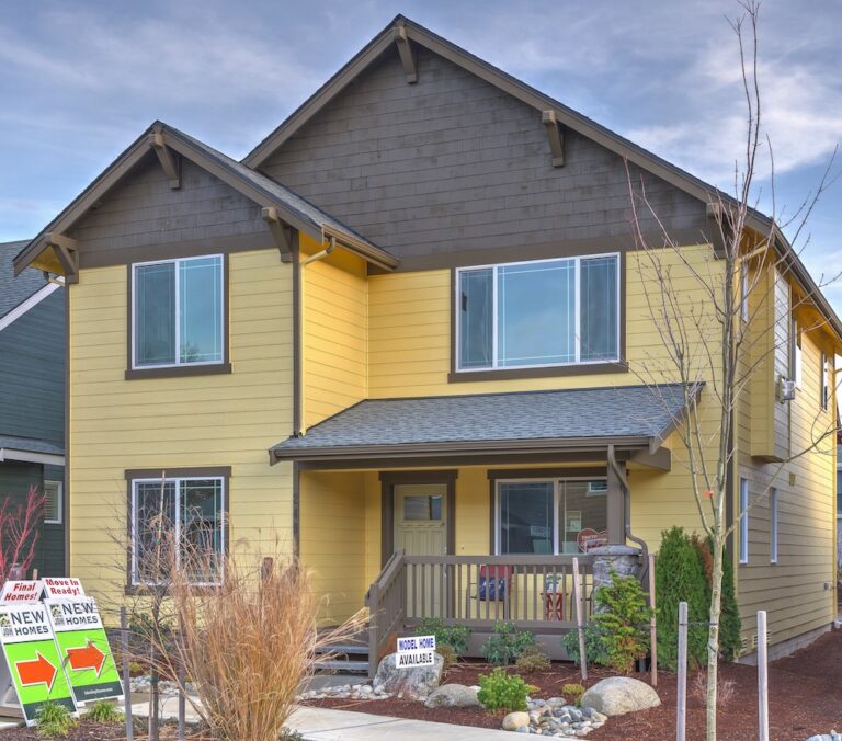  A two-story yellow house with a gray roof, featuring a front porch and multiple windows, has real estate signs indicating new homes available. 