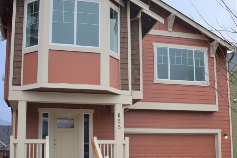 This is a two-story house with a beige and red exterior, a protruding bay window, a garage, steps leading up to a porch, and some landscaping.