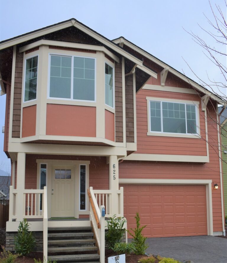  A two-story house with a beige and reddish-brown color scheme, a large front window, a garage, and steps leading to a covered porch. 