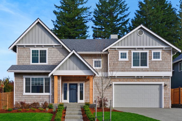 A two-story suburban house with shingle siding, a garage, and a gabled roof, surrounded by a well-manicured lawn and a clear blue sky.