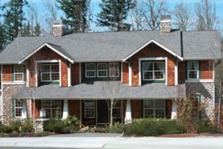 This is a two-story house with a gray shingle roof, stone accents, and dormer windows, surrounded by trees, without visible people.