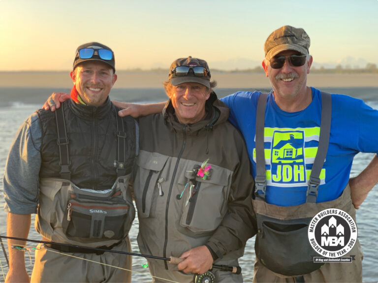 Three people stand on a beach at sunset, smiling with fishing gear, embracing each other. They are wearing casual attire with suspenders and caps.