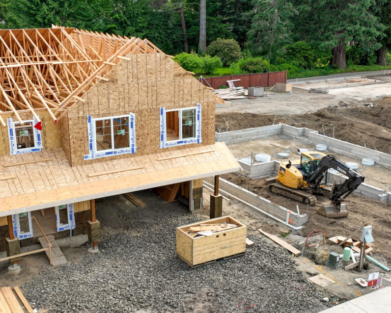 This aerial image shows a residential construction site with an unfinished wooden house frame featuring exposed beams, surrounded by dirt and building materials.