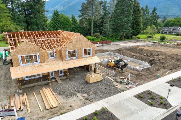 Aerial view of a residential construction site with a partially built wooden house frame, workers, scattered lumber, heavy machinery, and forested background.