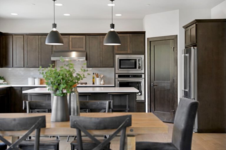 Modern kitchen with dark cabinetry, stainless steel appliances, white countertops, an island with seating, and pendant lighting over a wooden dining table.