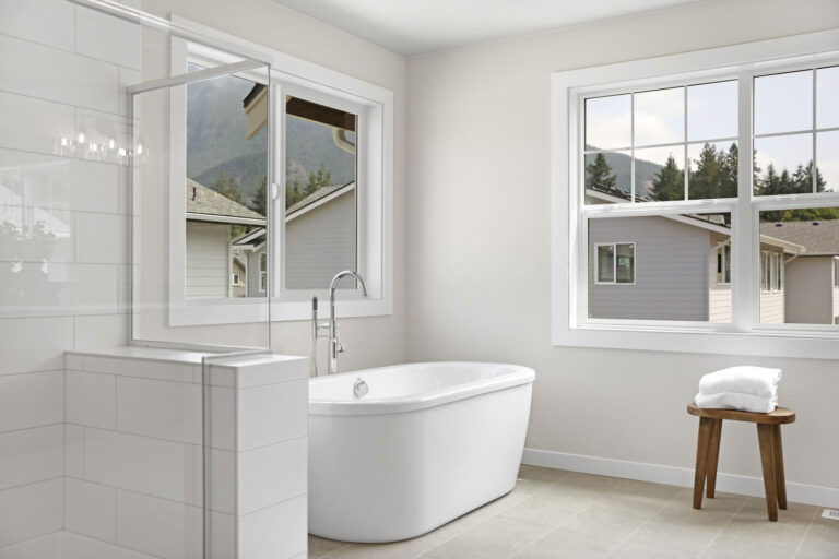 A modern bathroom with a freestanding tub, tiled walls, a small wooden stool with towels, and two windows offering a view of mountains and houses.