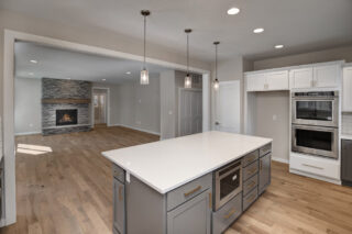 A modern kitchen with white countertops, gray cabinets, and stainless-steel appliances opens into a living area with a stone fireplace and hardwood floors.