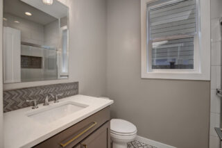 This image shows a modern bathroom with gray walls, a large mirror, a white vanity with cabinets, a sink, a toilet, and a glass-door shower.