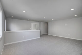 This is an empty, well-lit room with gray walls, carpet flooring, recessed ceiling lights, a half-wall ledge, and a doorway leading to another room.