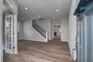 A modern interior hall with wooden flooring, a staircase with a black railing, white walls, recessed lighting, and an open French door.