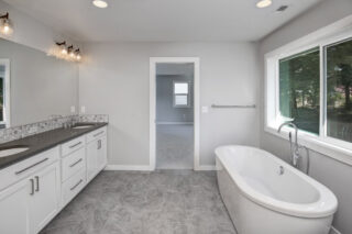 Modern bathroom interior with dual sinks, white cabinetry, freestanding bathtub, gray tiles, large window with a view of trees, and neutral colors.