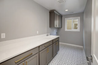 Modern laundry room with gray cabinetry, white countertop, patterned tile floor, and a small window with blinds. The space is clean and uncluttered.