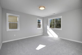 Empty room with gray carpet, white walls, two windows with blinds, some sunlight shining through, and a flush mount ceiling light fixture.